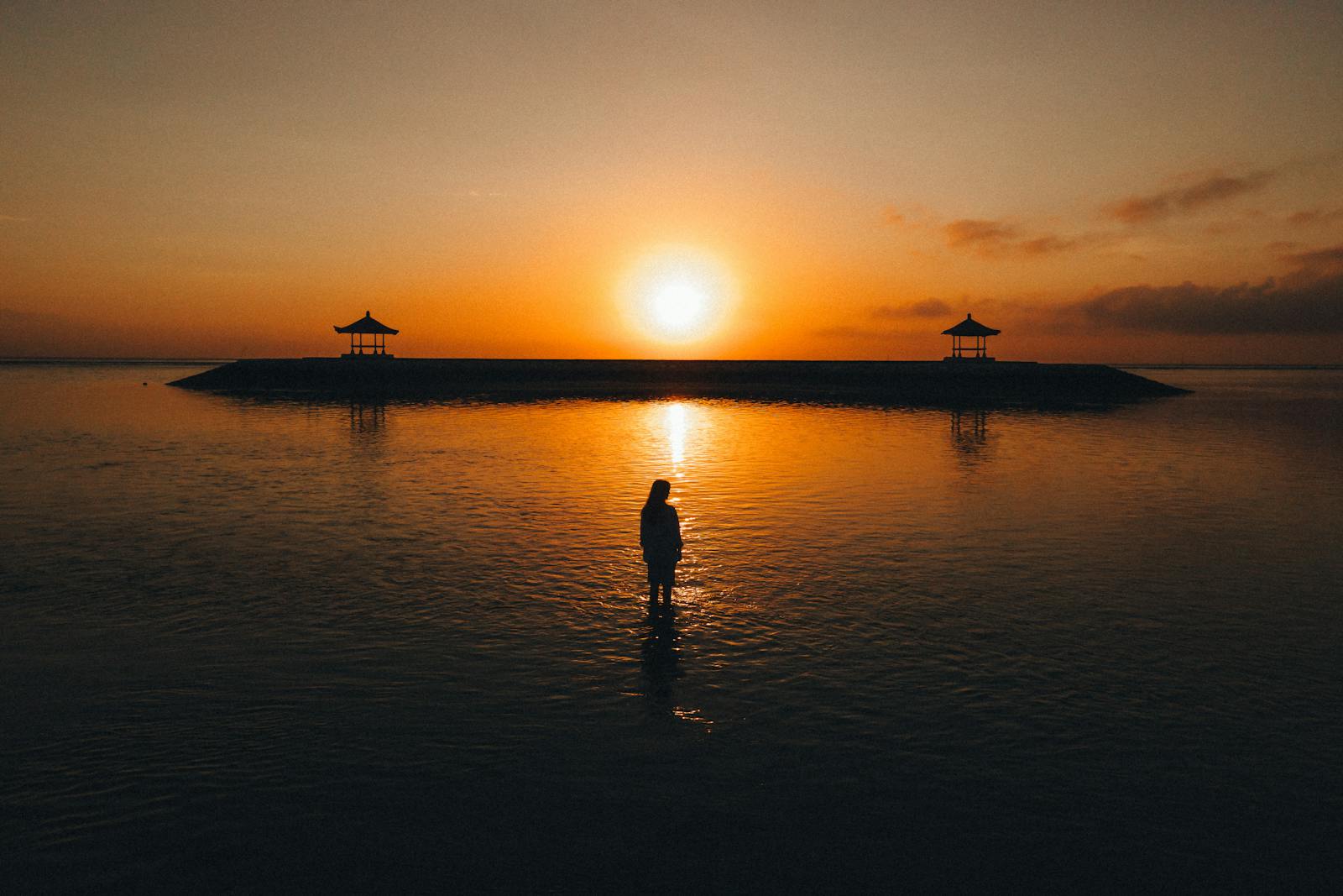 A tranquil scene of a silhouette at sunrise near pavilions on a Bali beach.